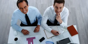 Two accountants Financial Controller and a Bookkeeper at a desk with laptop, PC, diaries, charts, and coffee, collaborating on small business finances.