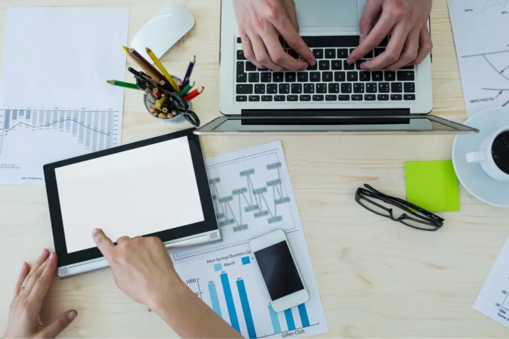 An overhead view of a modern workspace with a person using a tablet and laptop to manage financial charts, illustrating what is digital bookkeeping in a paperless office.