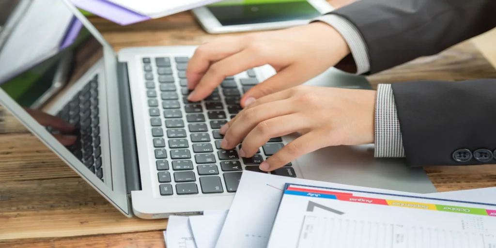 Business professional typing on a laptop while reviewing financial documents at a desk.