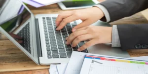 Business professional typing on a laptop while reviewing financial documents at a desk.