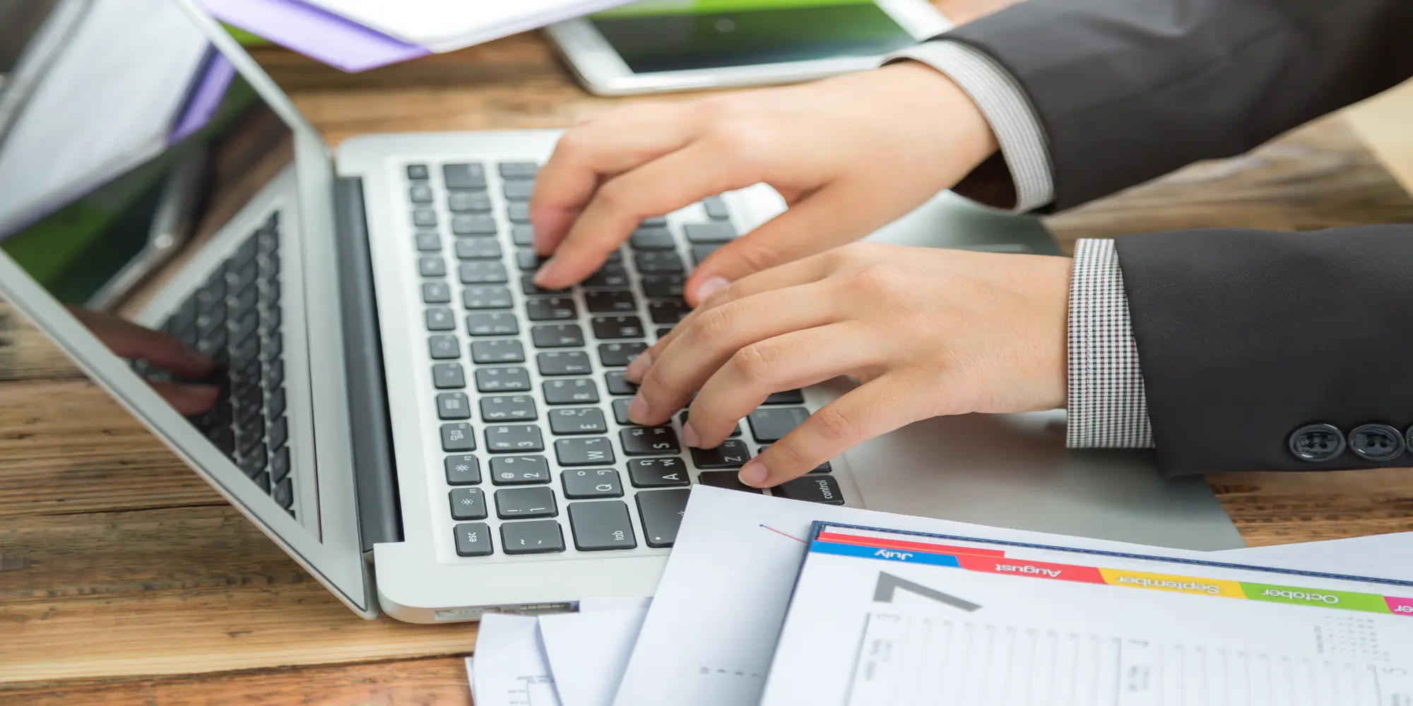 Business professional typing on a laptop while reviewing financial documents at a desk.
