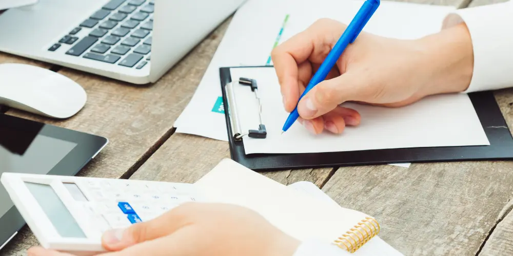 Accountant holding a calculator and writing financial journal entries on a clipboard at a desk with a laptop.